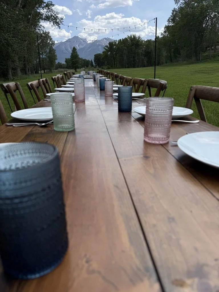 Long outdoor dining table set with plates, colorful glasses, and chairs, outdoors in a grassy area with trees and mountains in the background, under string lights.