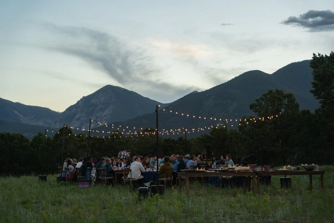 People gathered at an outdoor evening event with string lights, mountains in the background, and a partly cloudy sky.
