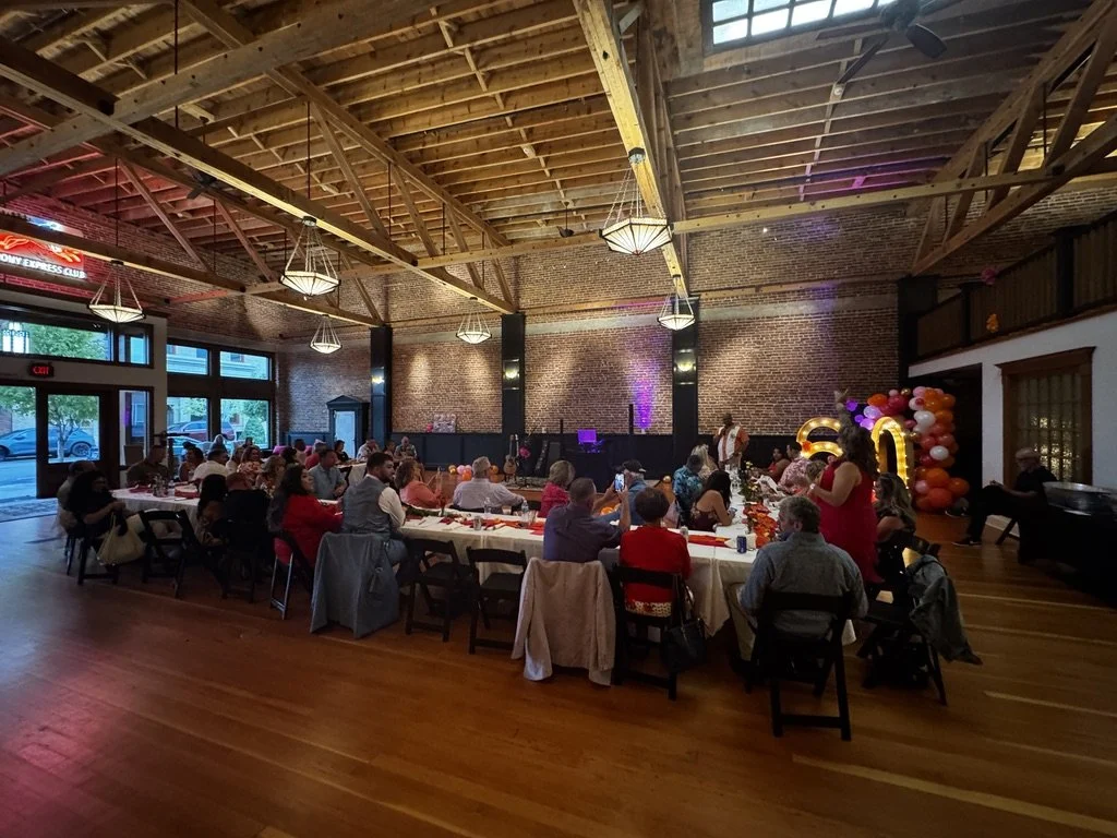 A large gathering of people seated at long banquet tables inside a building with wooden beams, brick walls, and large windows. The event appears to be a celebration with decorations, including balloons and large illuminated numbers.