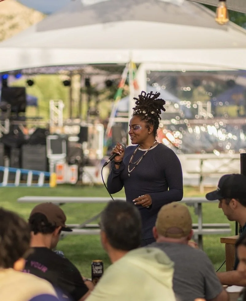 A woman with dreadlocks styled in a tall updo, wearing a black long-sleeve top and silver jewelry, speaking into a microphone at an outdoor event under a covered pavilion, with a blurred crowd and scenic background.