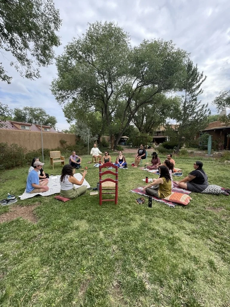 A group of people sitting on blankets and cushions in a circle outdoors in a grassy yard, under a tree, with houses in the background.