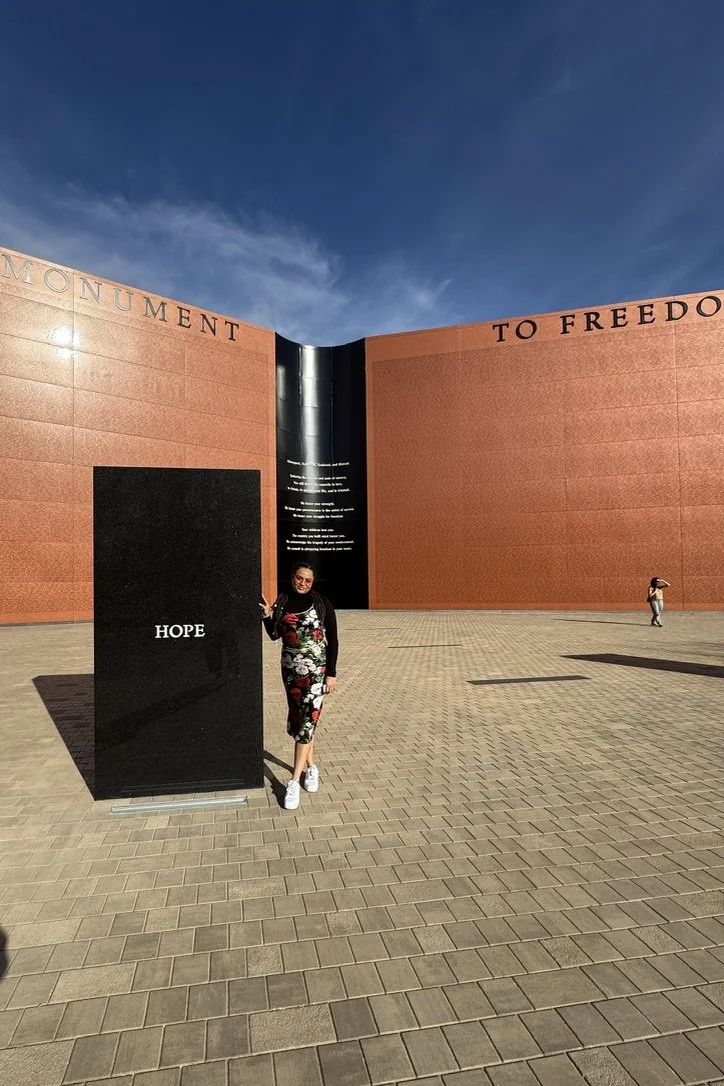 A woman standing next to a black stone monument with the word "HOPE" inscribed, in front of a large memorial wall with inscriptions. The scene is outdoors under a blue sky with some clouds, and the ground is paved with brick-like tiles.