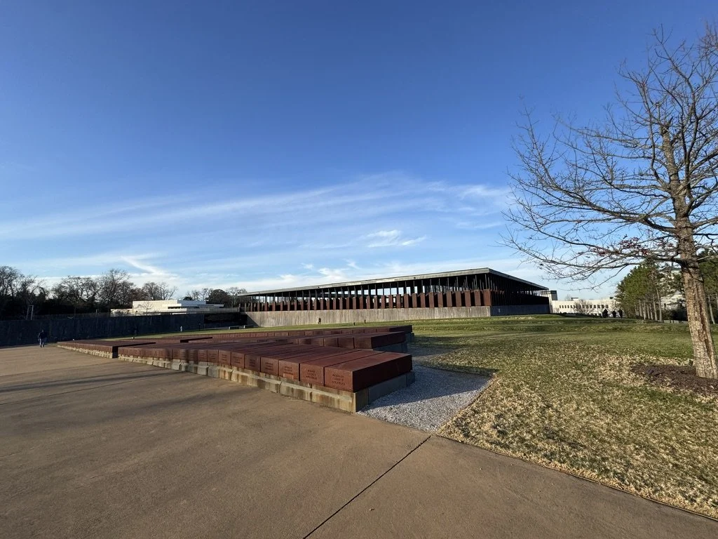 A large outdoor seating area with rust-colored benches on a concrete platform, a grassy slope with a lone leafless tree, and a modern building with a sloped roof in the background under a clear blue sky.