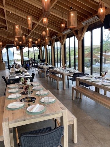 Empty restaurant with wooden tables and chairs, set for dining, with large windows providing a view of greenery outside, and warm pendant lighting hanging from the wooden ceiling.