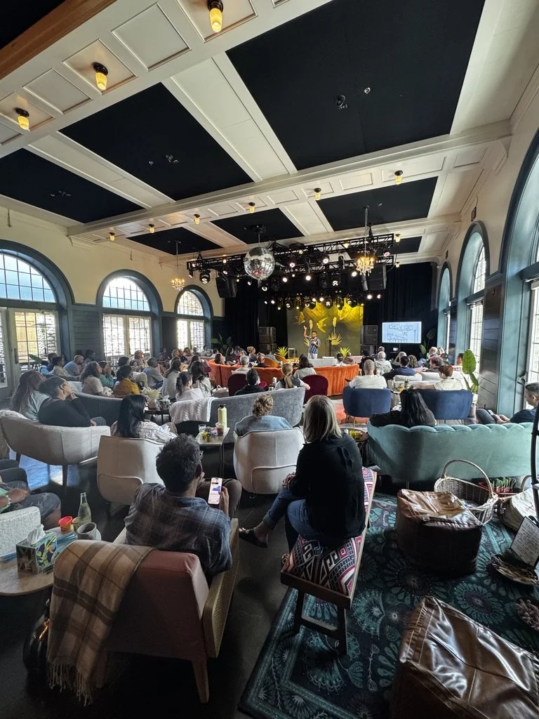 A large indoor event space with high arched windows, decorating the walls, and a stage at the front where a presenter is speaking. The room is filled with people seated in various upholstered chairs and sofas, listening to the presentation. There are