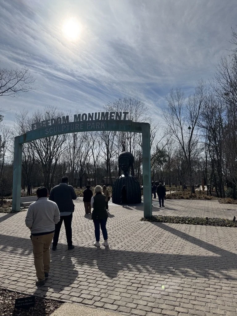 People walking towards the entrance of Freedom Monument Sculpture Park, which features a large bust sculpture and an archway with park name; leafless trees and a cloudy sky visible.