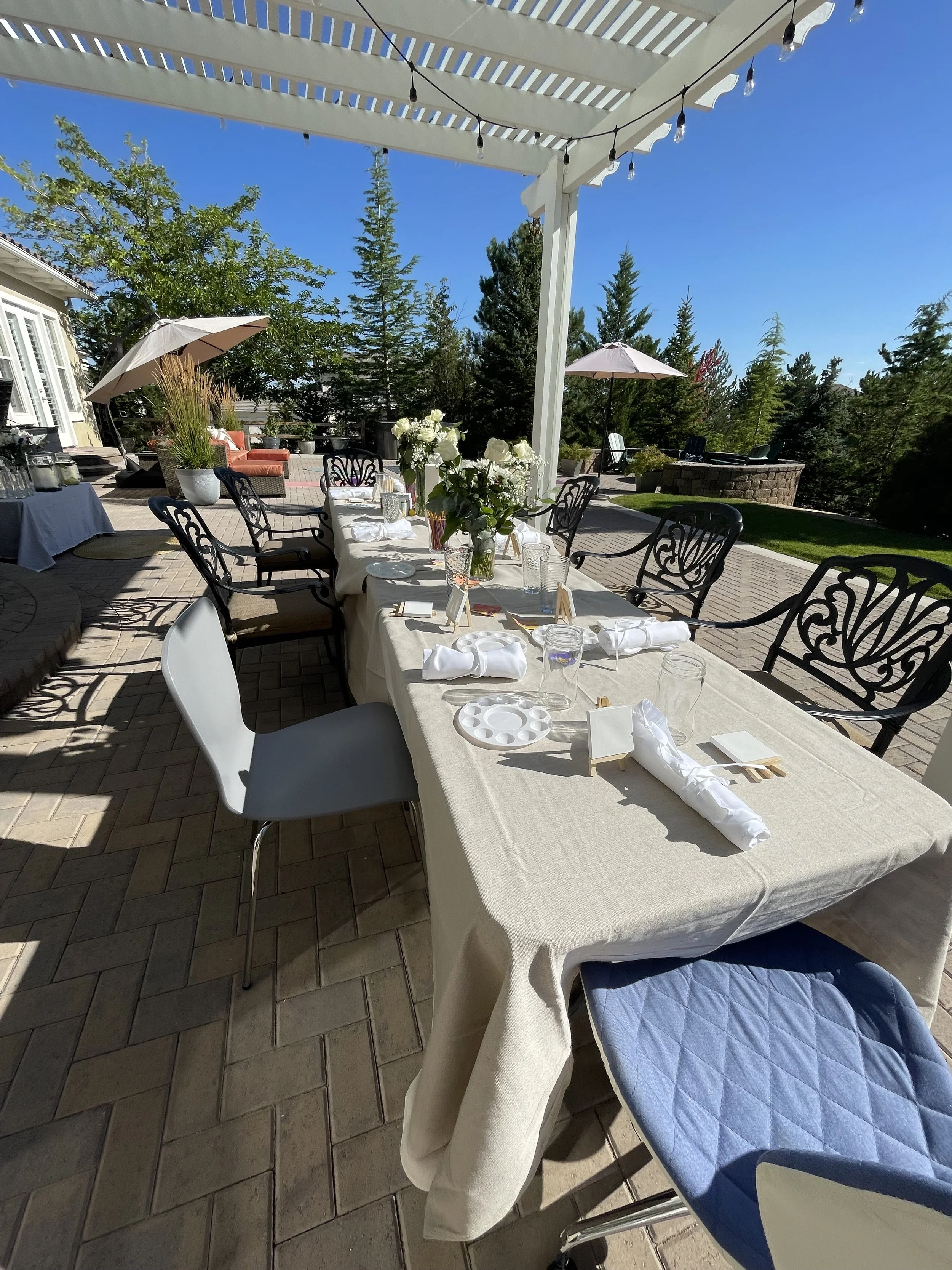 Outdoor dining setup for wellness or community workshop celebration in Nevada – table with white tablecloth, floral centerpieces, plates, glasses, and small easels under pergola with string lights, facilitated by ALIGN Agency