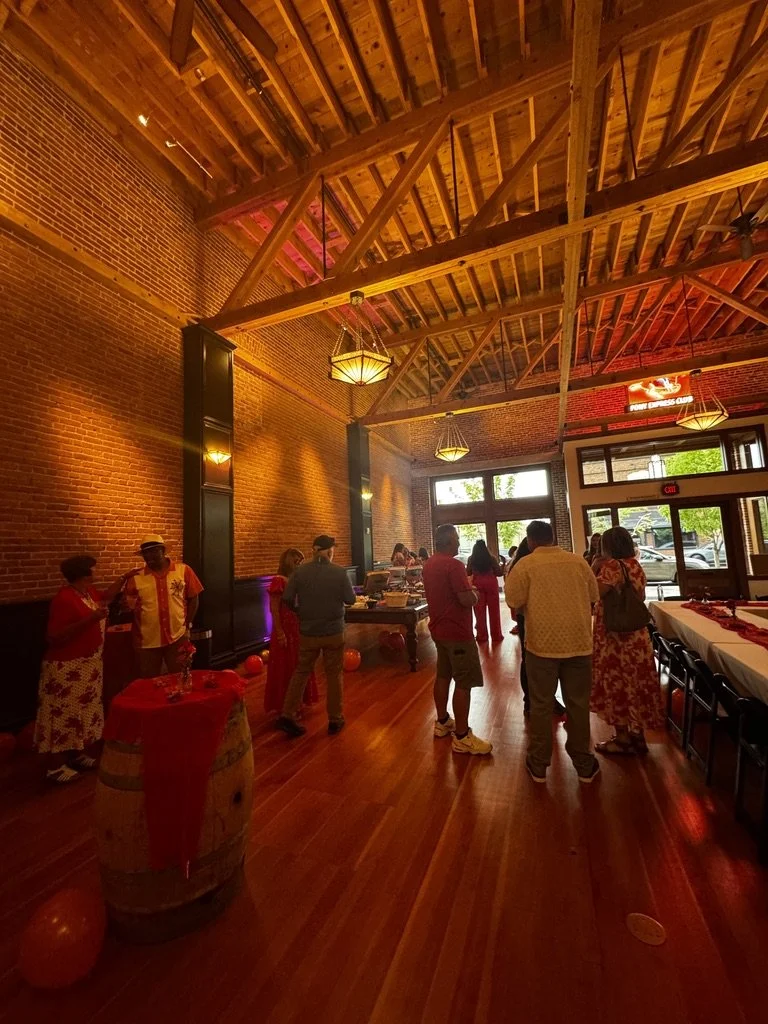 People gathered inside a restaurant with brick walls and a wooden ceiling, decorated with festive balloons and table settings, possibly for a celebration or party.