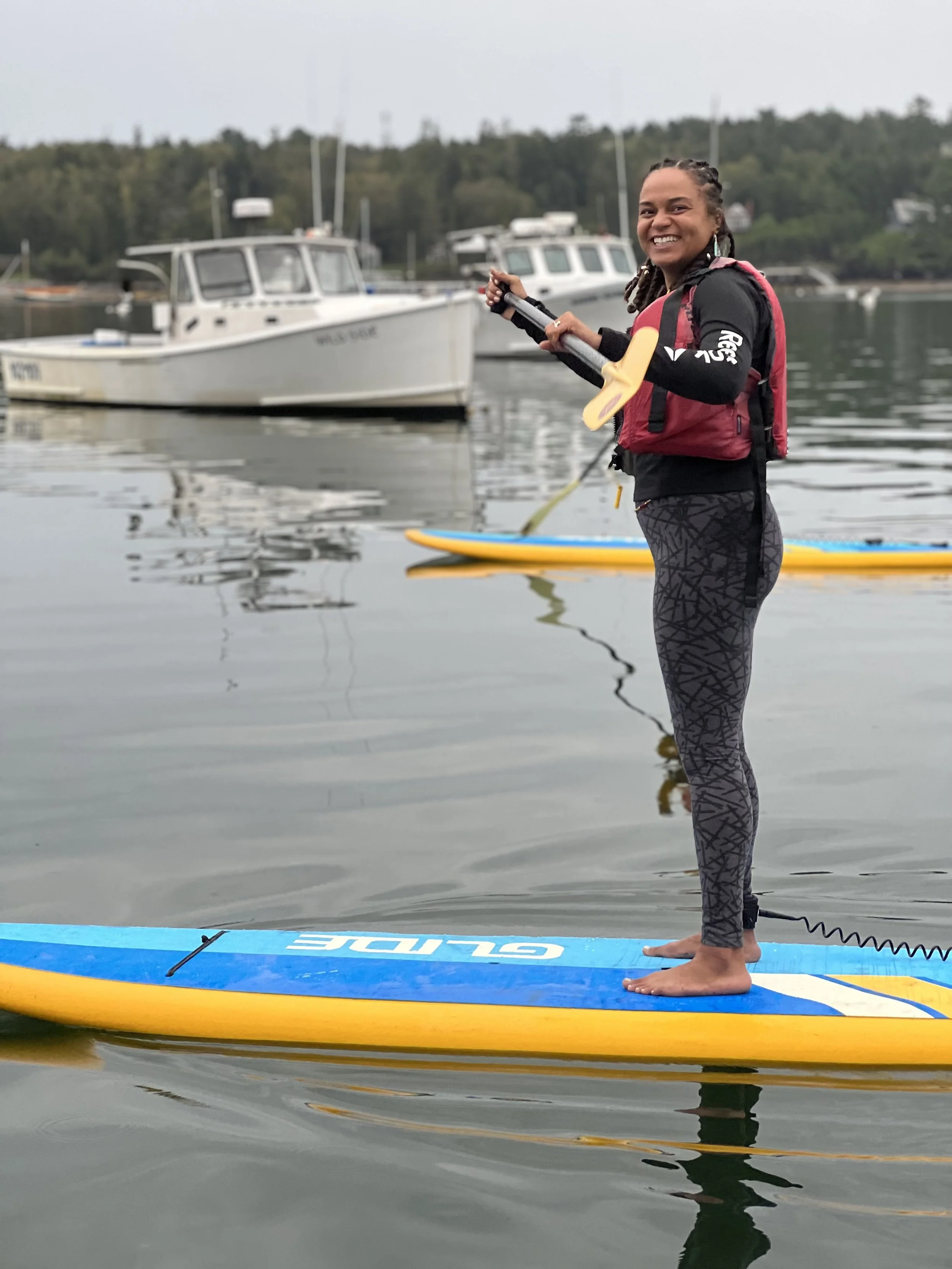 Outdoor wellness or retreat workshop for mission-driven organizations in Nevada – woman standing on paddleboard on calm water, smiling, with boats and tree-lined shoreline in background, facilitated by ALIGN Agency