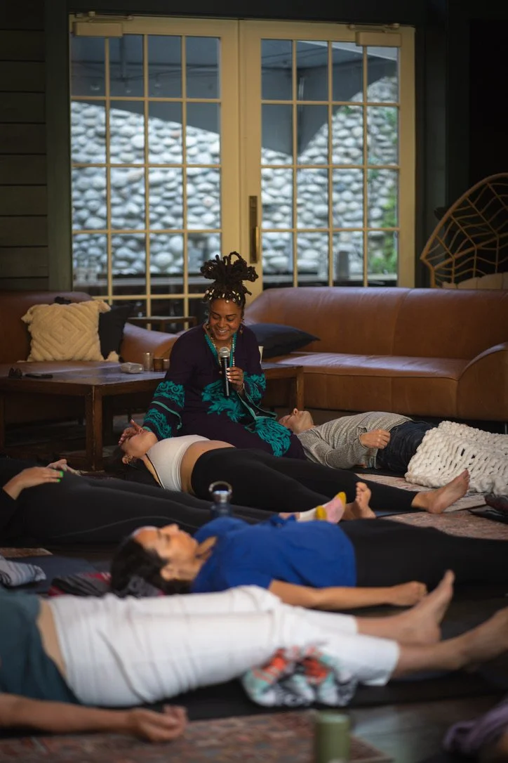 A woman with dreadlocks holding a microphone, sitting on the floor, surrounded by people lying on yoga mats in a cozy room with large windows and a stone wall outside.