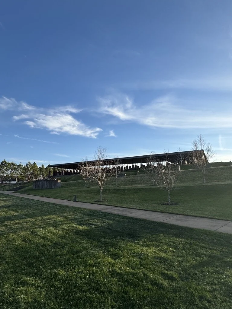 A grassy area with a sidewalk, a few small leafless trees, and a modern building with a covered walkway underneath, set against a bright blue sky with some clouds.