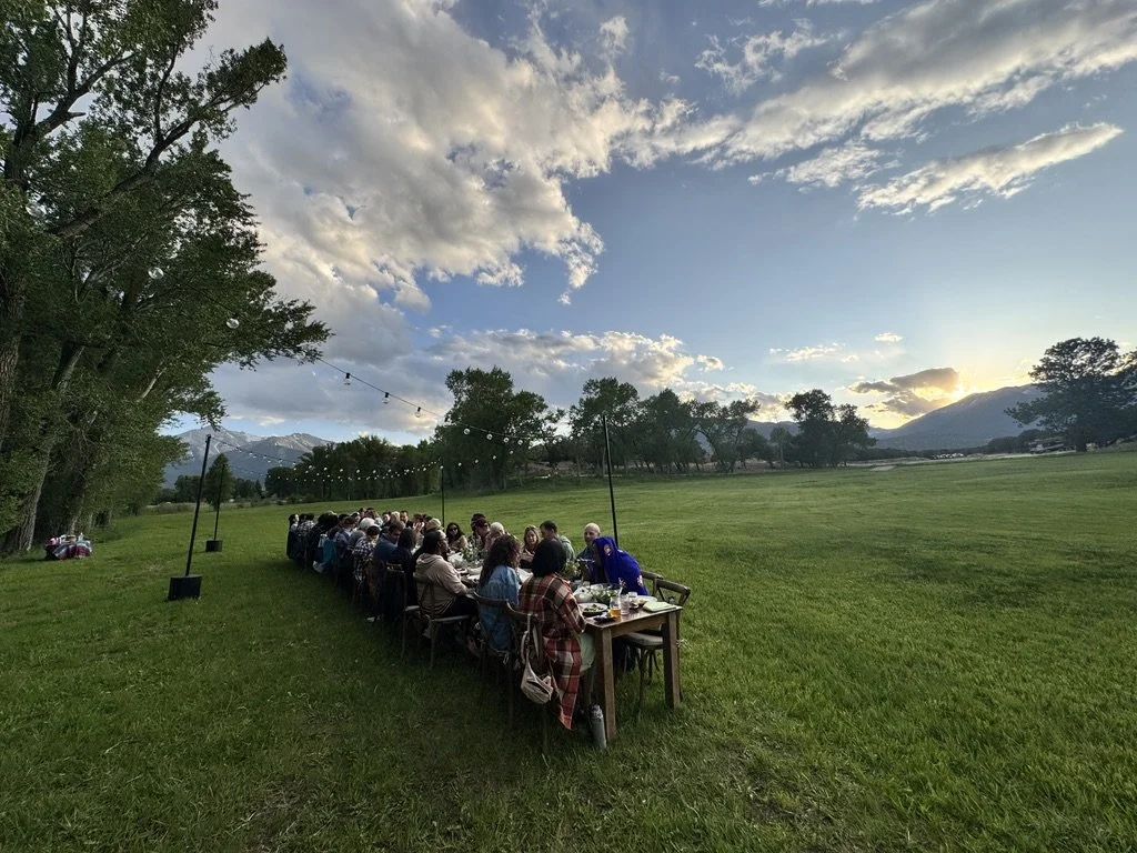 Outdoor gathering with many people sitting at a long table in a grassy field during sunset, with string lights overhead and mountains in the background.