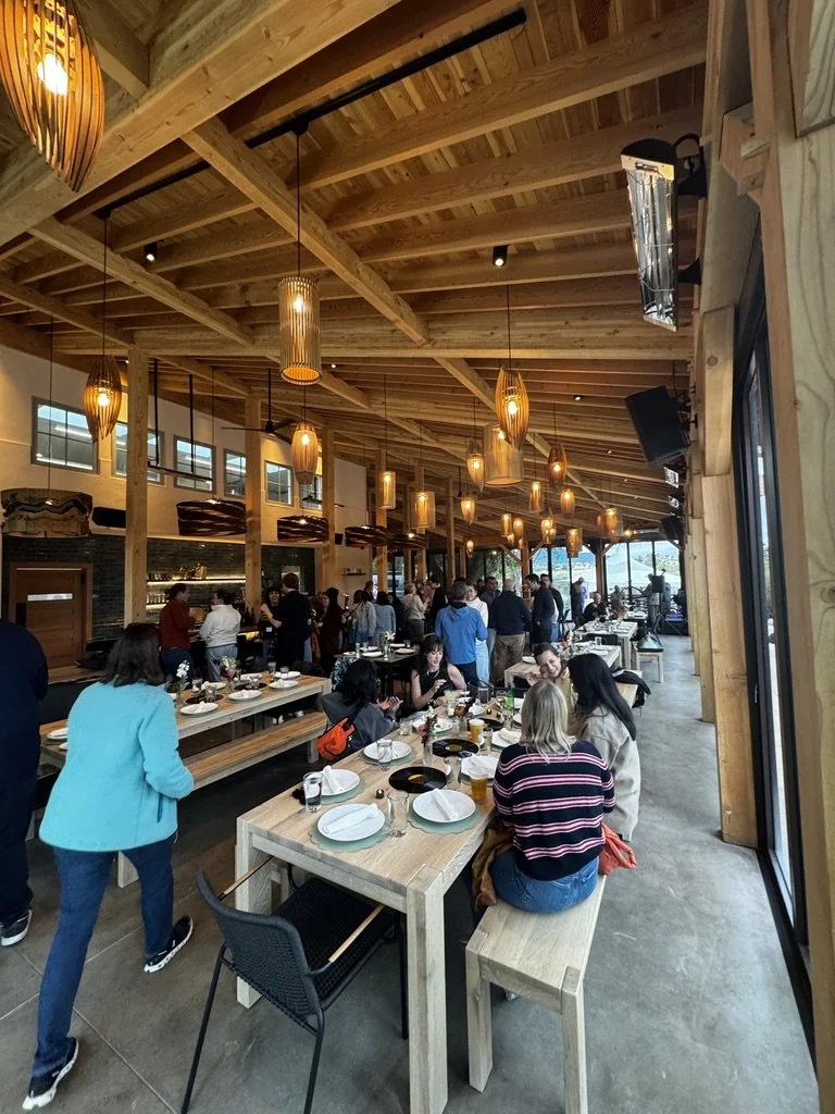 Interior of a busy restaurant with wooden beams and hanging lights. People are seated at tables and waiting in line, with a bar or counter area visible in the background.
