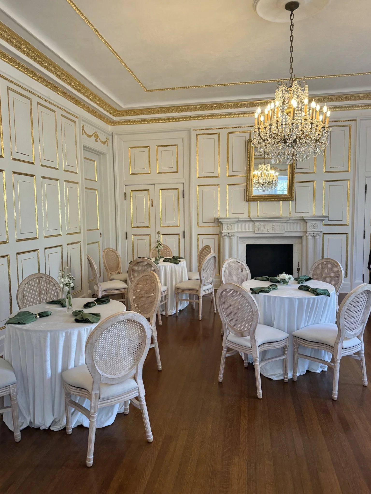 Elegant dining room with white walls decorated with gold trim, a marble fireplace, a chandelier, and round tables with white tablecloths, green napkins, and chairs.