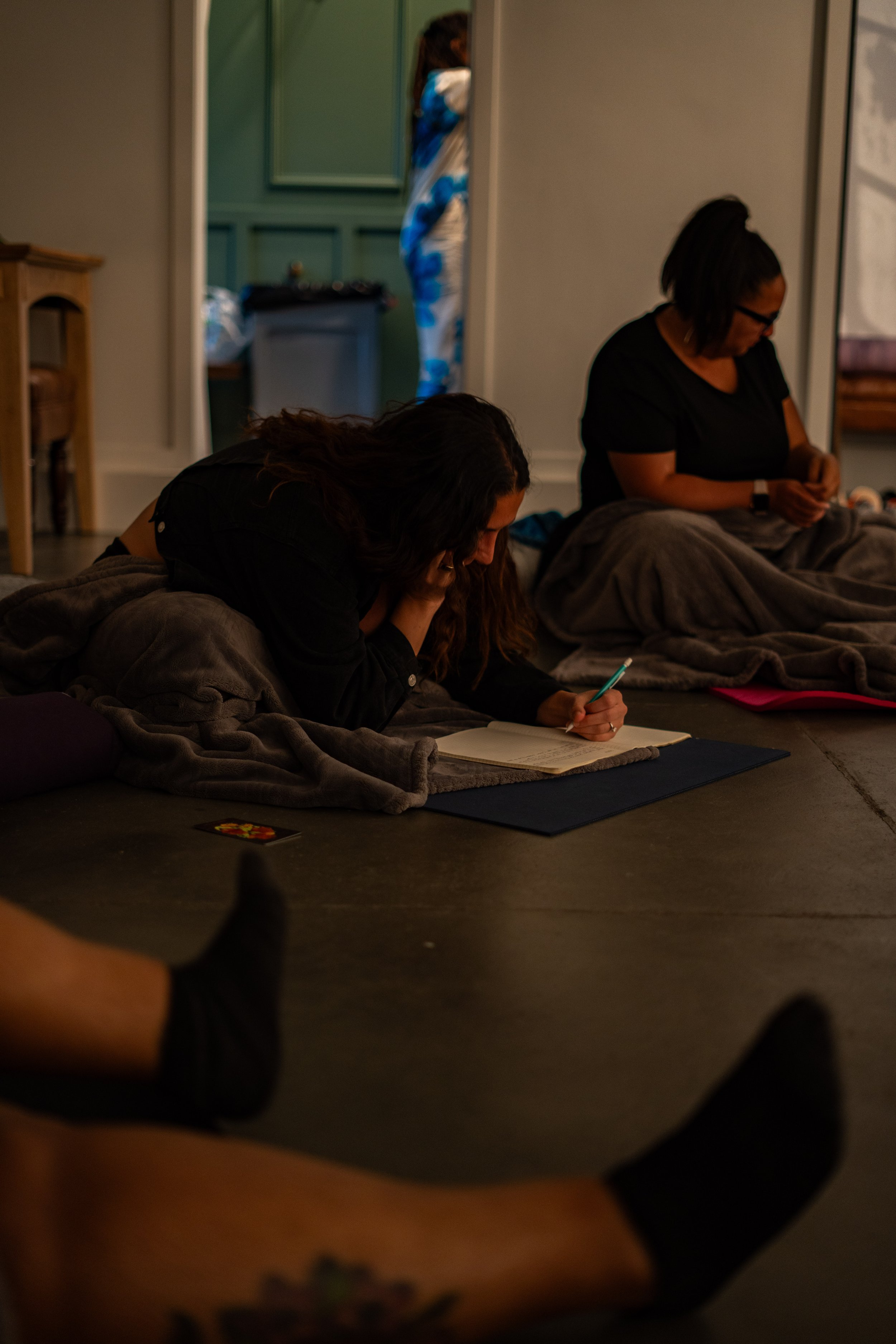 Indoor wellness or creative workshop for mission-driven organizations in Nevada – two women sitting on floor over blankets, writing or drawing in notebooks in dimly lit room, with another participant in background, facilitated by ALIGN Agency