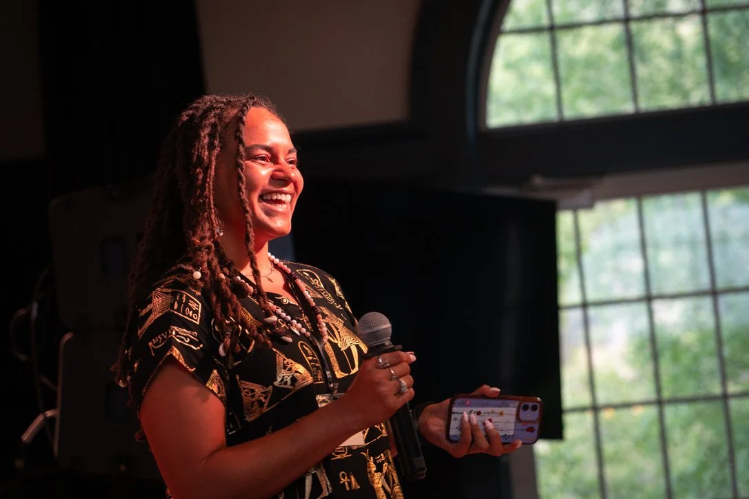 A woman with dreadlocks holding a microphone and a smartphone, smiling while speaking at an indoor event with large windows showing trees outside.