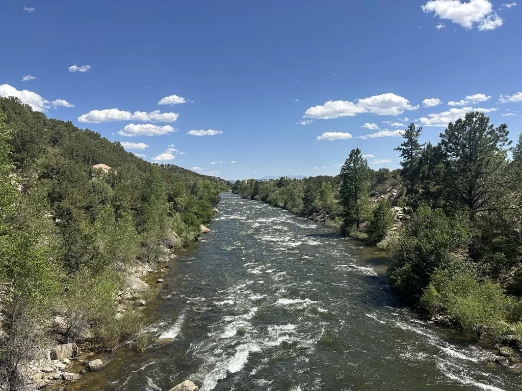 A scenic view of a river flowing through a lush green landscape under a blue sky with scattered white clouds.