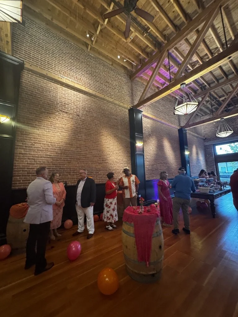 People in semi-formal attire gather indoors at a party or celebration, with balloons on the wooden floor and a decorated barrel in the foreground. The space has an exposed brick wall, wooden ceiling beams, and hanging light fixtures.