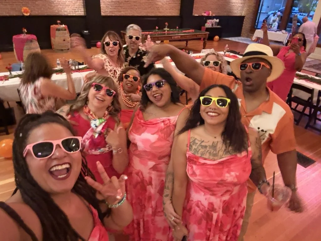 A group of people at a party, smiling and wearing colorful sunglasses and pink or red outfits, enjoying themselves in a decorated indoor setting.