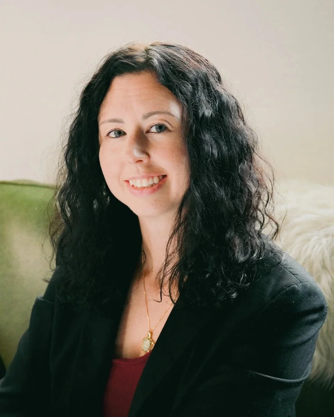 A woman with curly black hair and fair skin smiling, wearing a black blazer over a red top, and a gold necklace, sitting on a green couch with a light-colored background.