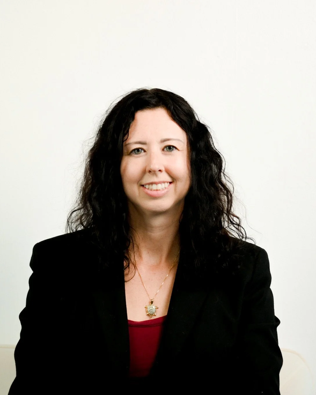 A woman with shoulder-length curly black hair smiling, wearing a black blazer and a necklace with a large decorative pendant, against a plain light-colored background.