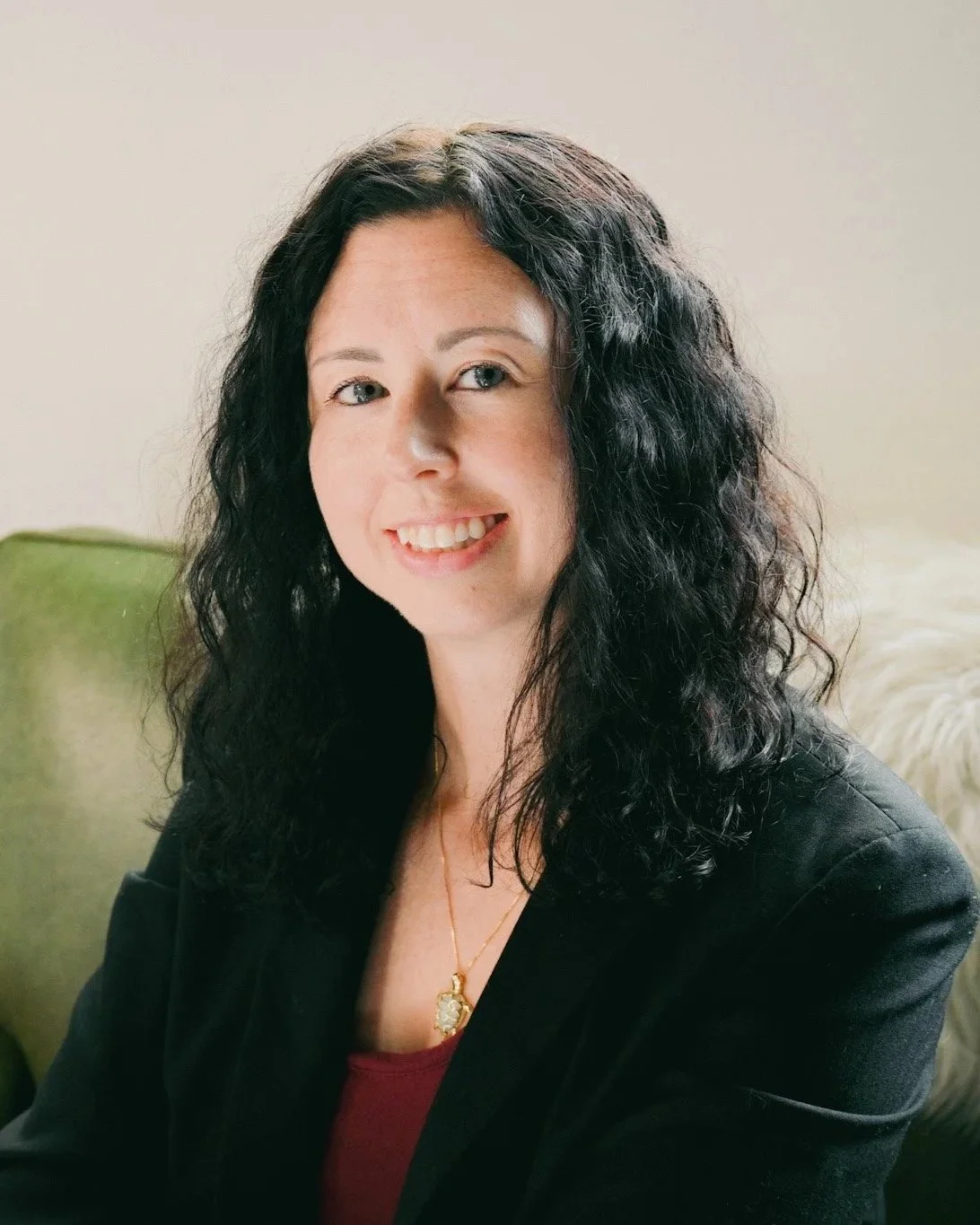 A woman with long black curly hair smiling, wearing a black blazer and a gold necklace, sitting on a green couch with a cream-colored wall behind her.