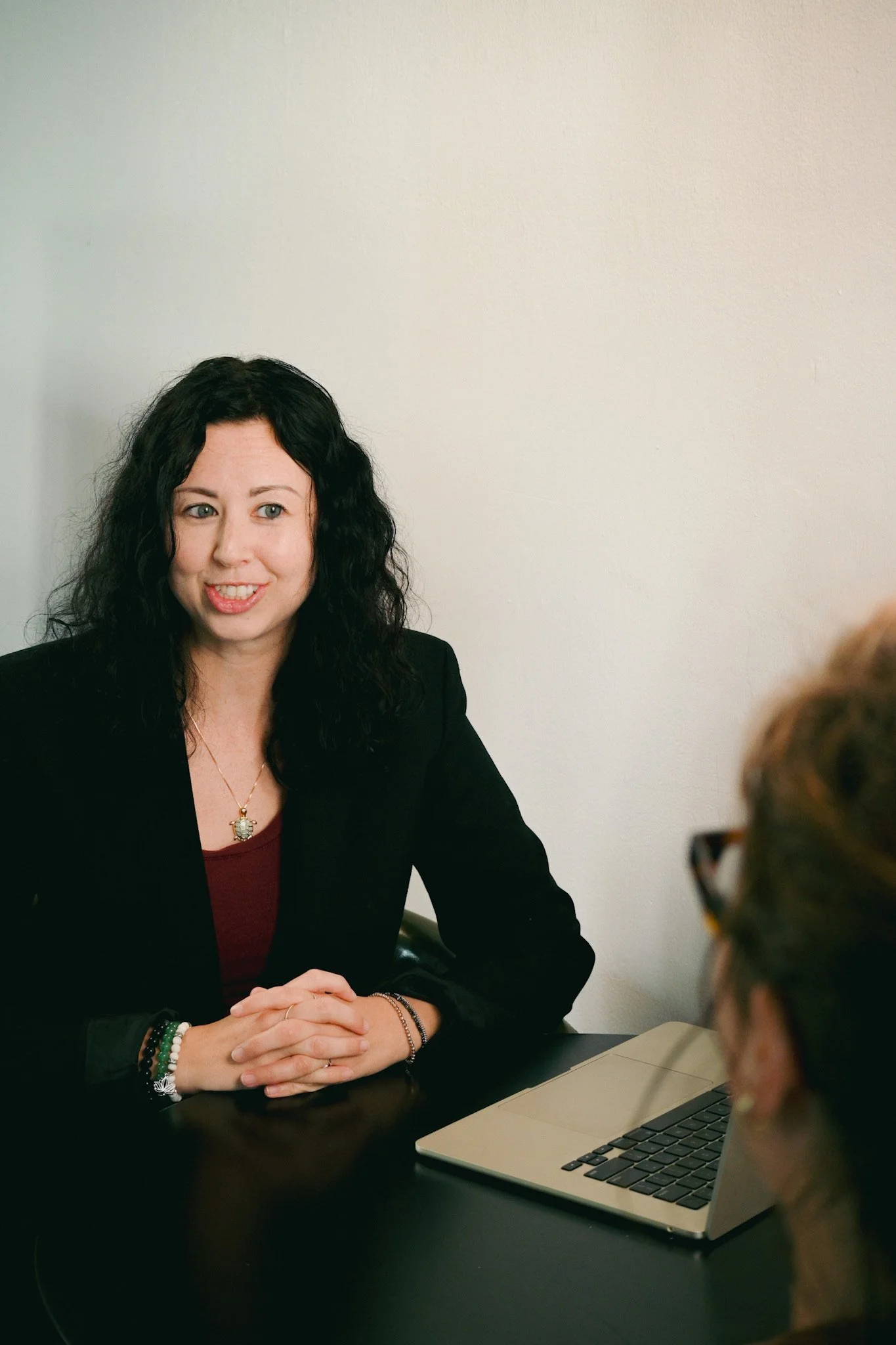 A woman with dark curly hair wearing a black blazer and red top, sitting at a table with a laptop, engaged in conversation with another person whose head and glasses are partially visible in the foreground.