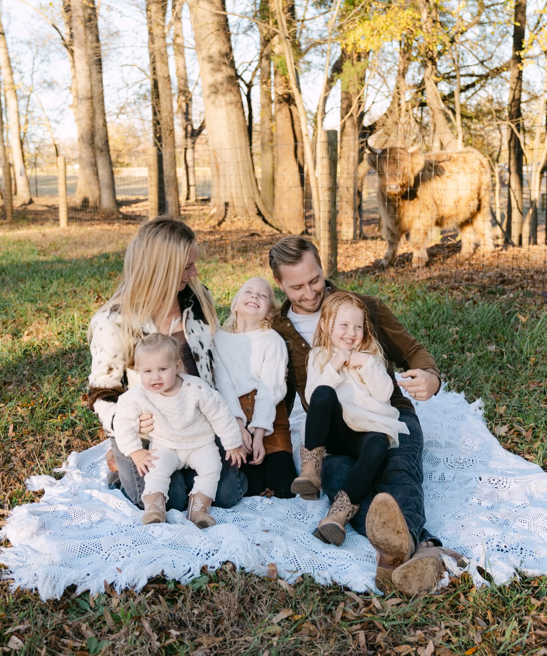 Family sitting on a blanket in a park, smiling, with a big furry animal behind the fence in the background.