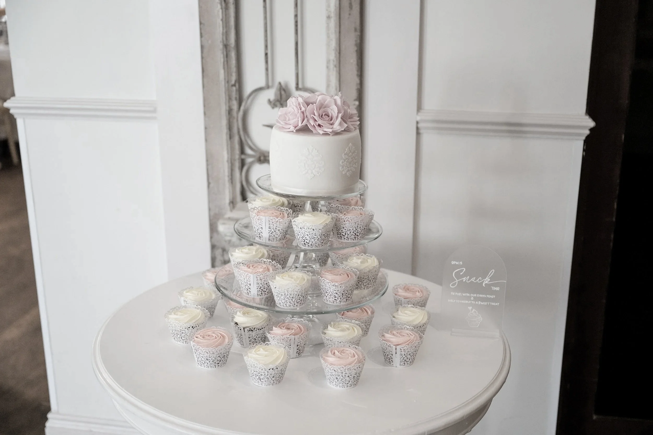 A white cake decorated with pink roses on top, displayed on a three tiered glass stand, surrounded by white and pink cupcake treats on a round white table near a white-paneled wall.