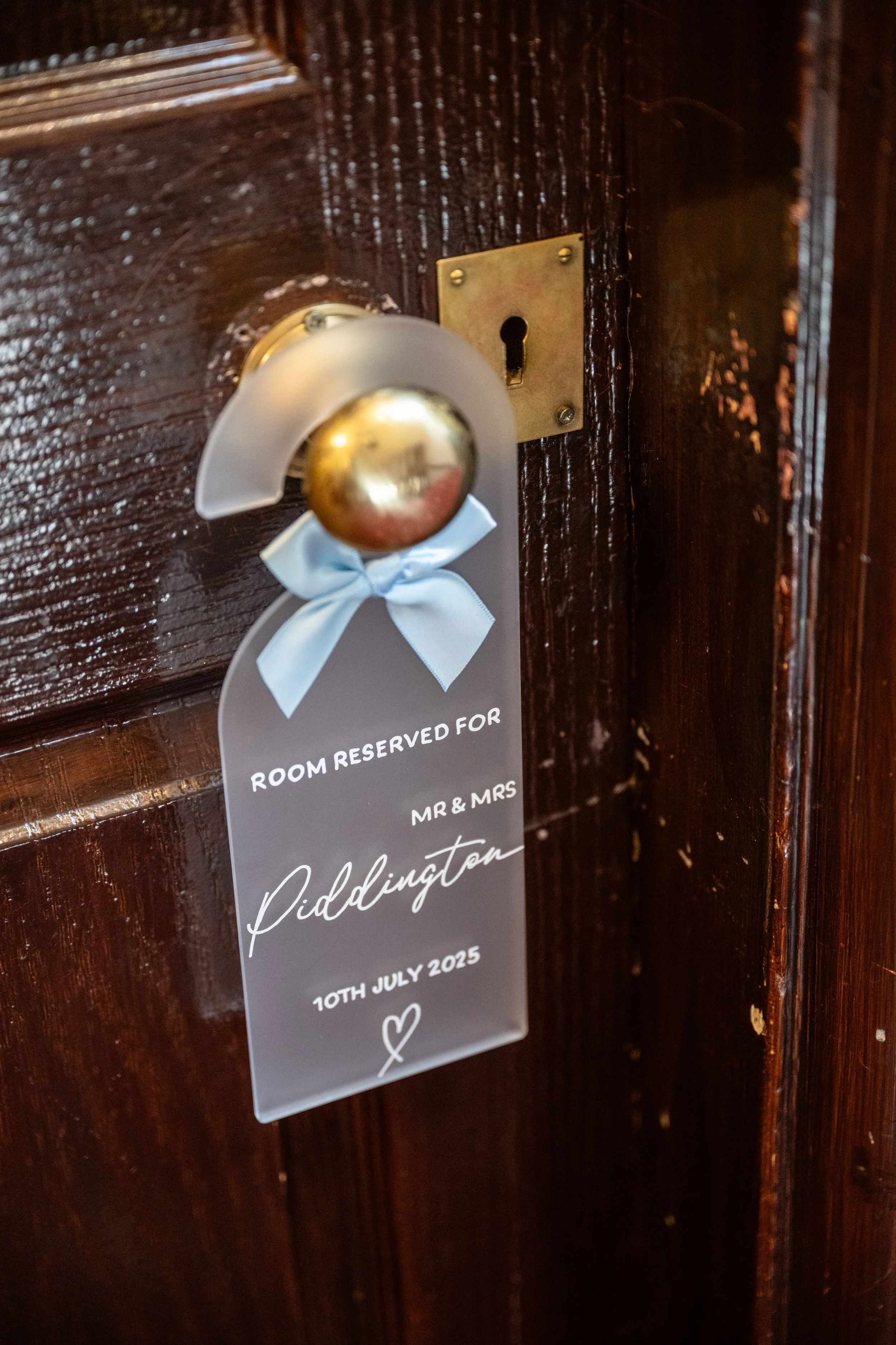 Door with reserved sign for Mr. and Mrs. Piddington, wedding date July 10th, 2025, attached to a shiny gold doorknob with a white ribbon. Personalised stationery for weddings and events.