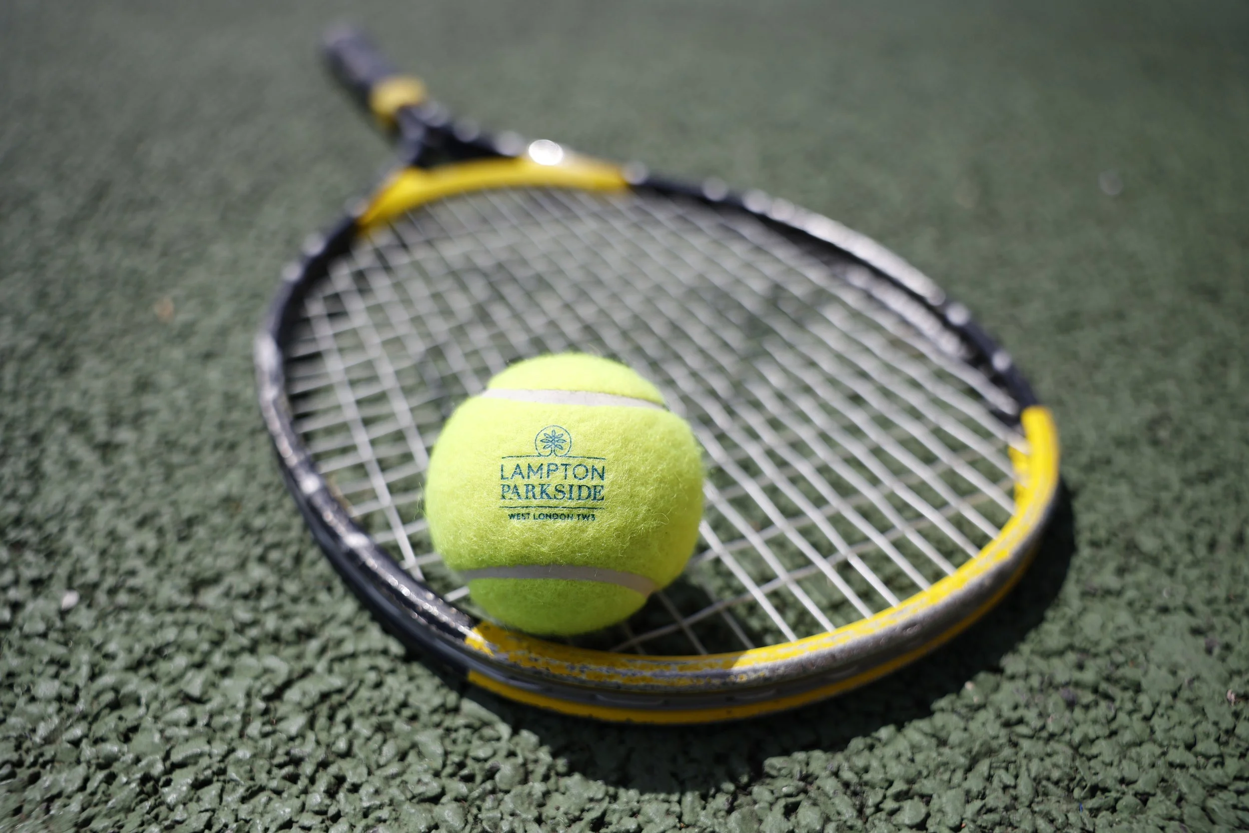 Tennis ball with the text 'Lampton Parkside West London TW3' resting on a tennis racket on a green tennis court. Personalised merchandise for corporate events.