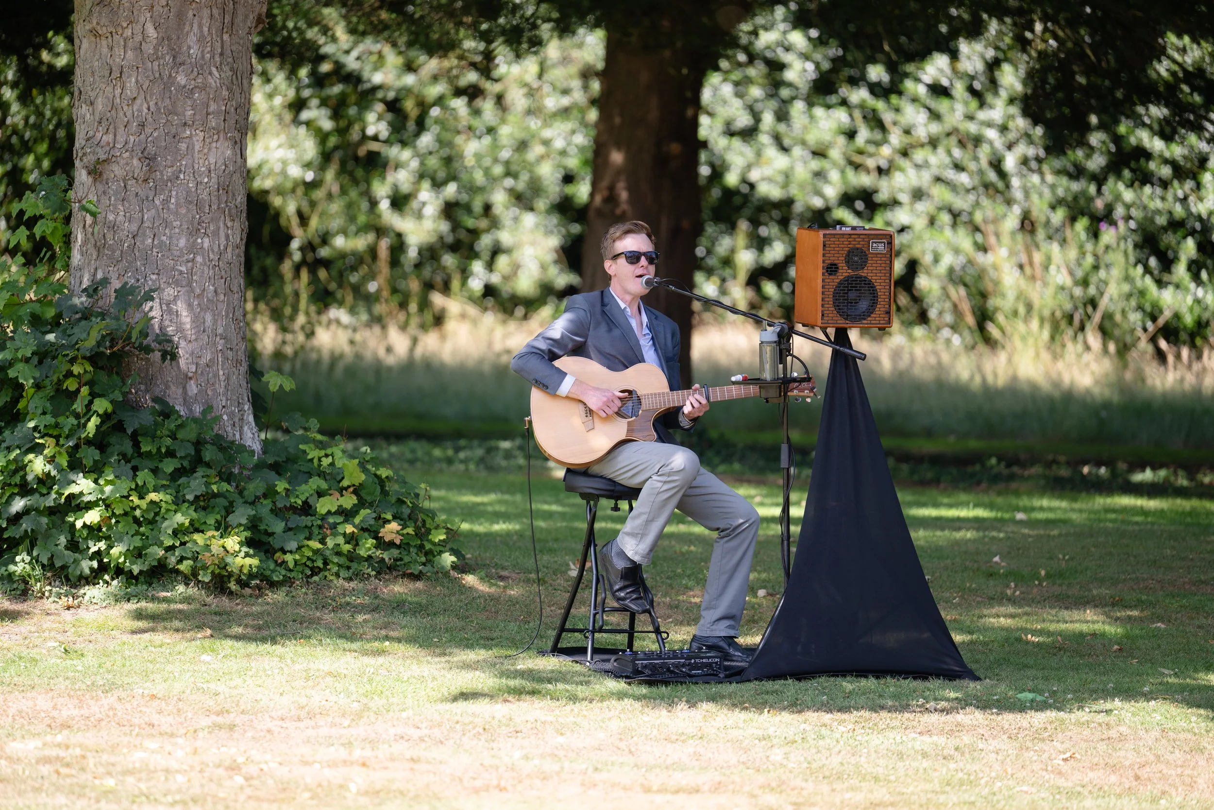 A man wearing sunglasses and a gray blazer playing acoustic guitar and singing into a microphone outdoors in a park with trees and grass.