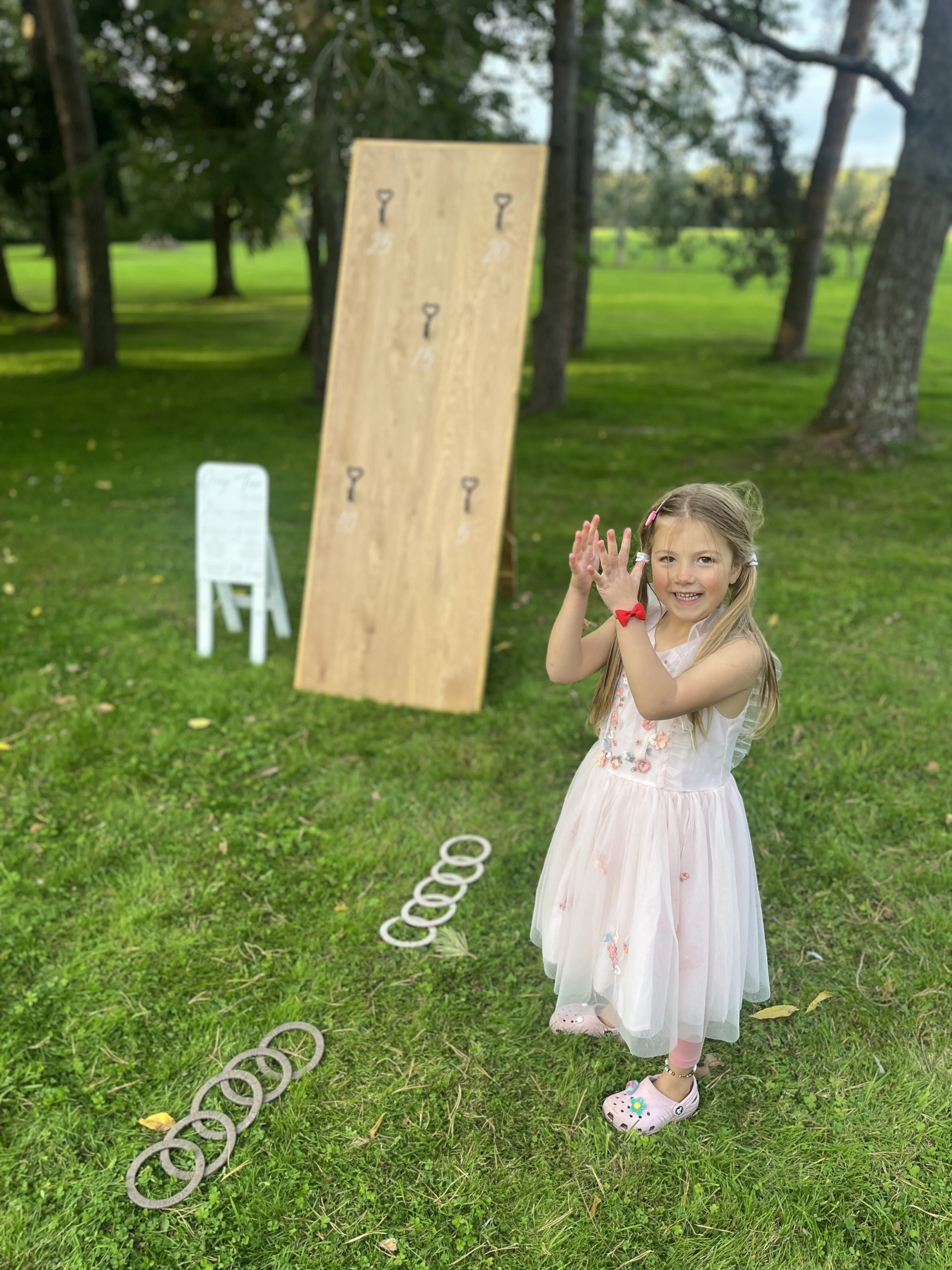 A young girl wearing a pink dress and crocs, clapping and smiling in an outdoor park with a ladder and rings set up for a game in the background. Ring Toss is part of a giant lawn games package for hire.