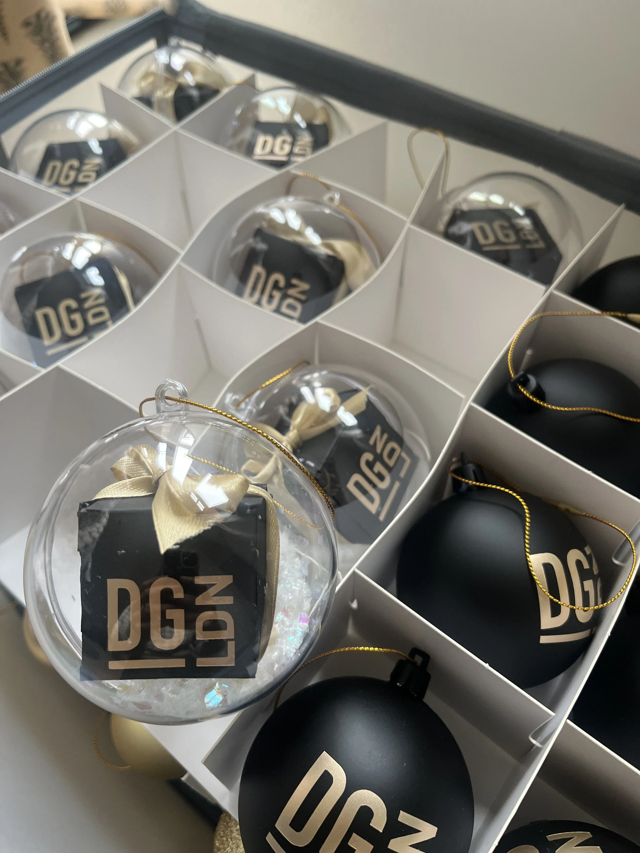 Assorted clear and black Christmas ornaments with gold accents, placed in individual compartments of a white display box. Personalised merchandise for gifts and events.