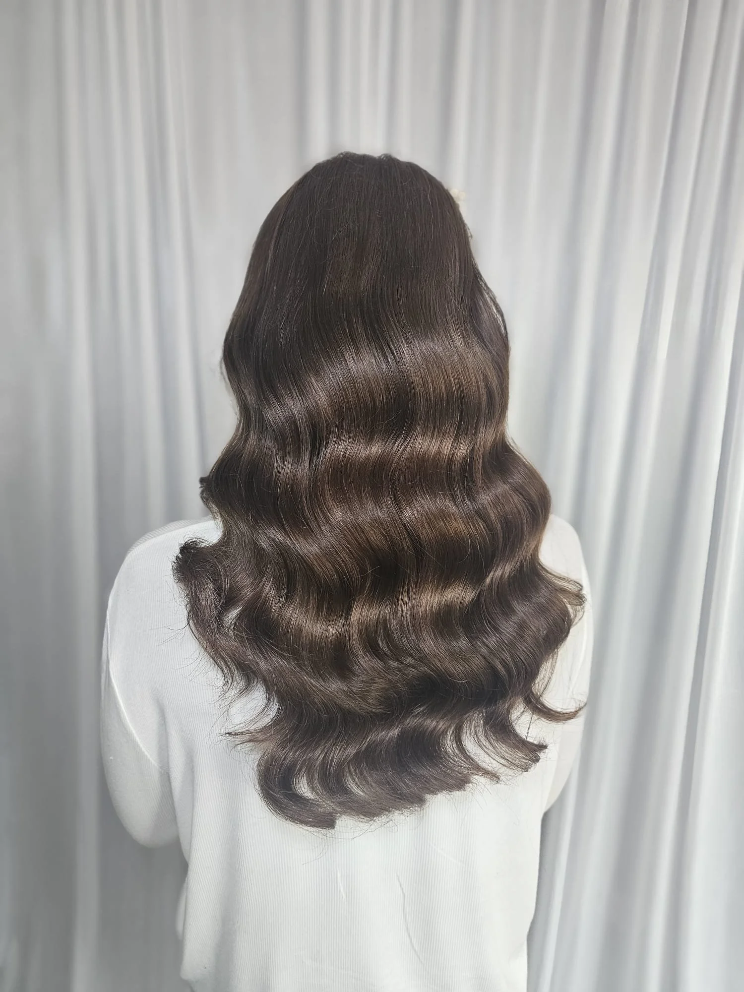 Back view of a person with long, dark brown, wavy hair standing in front of a gray curtain.
