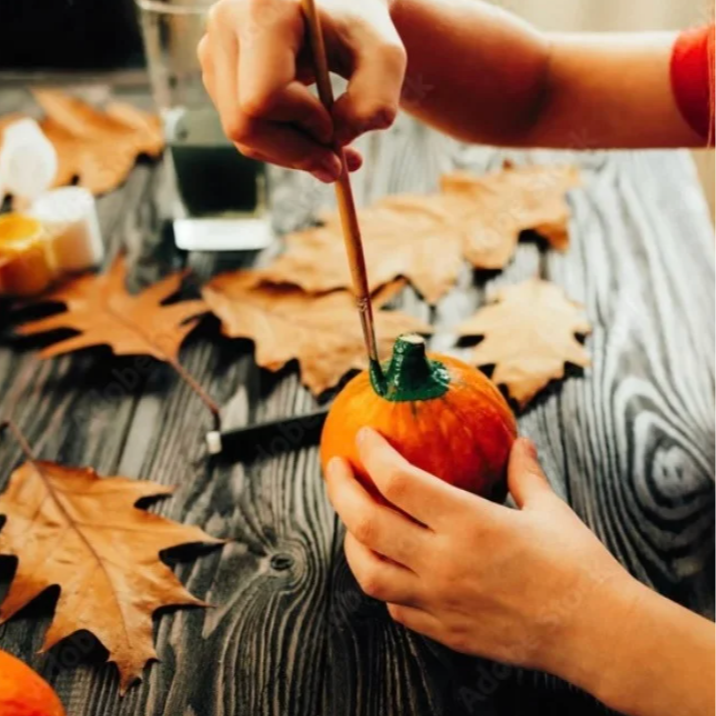 Person painting the top of a small pumpkin green with a brush, surrounded by large dried autumn leaves on a wooden surface.