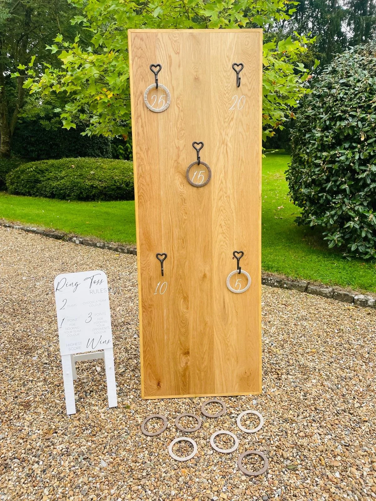 An outdoor ring toss game setup with a wooden board featuring five rings and numbered hooks, situated on a gravel ground with green trees and bushes in the background. Part of a giant lawn games package for hire.