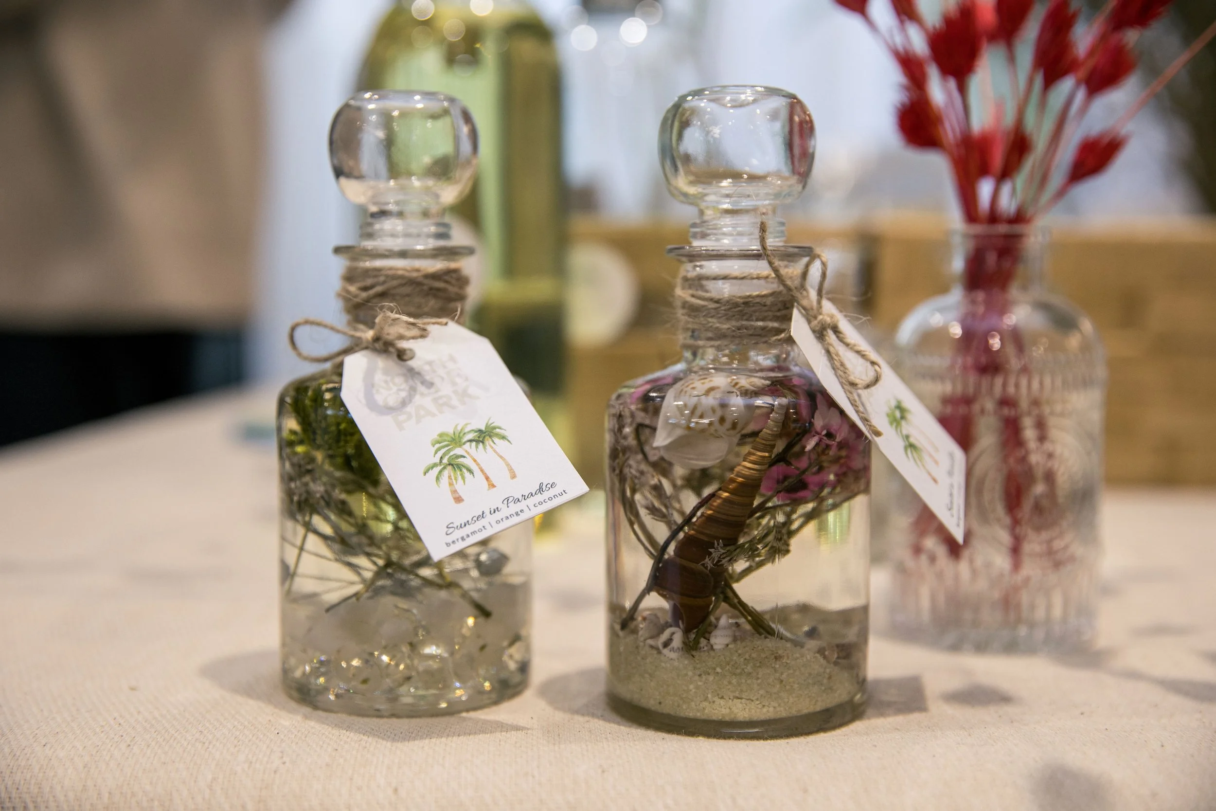 Three glass bottles filled with decorative items, adorned with twine and tags, on a light-colored surface with blurred background of flowers and more bottles. Themed event planning, for diffuser making.