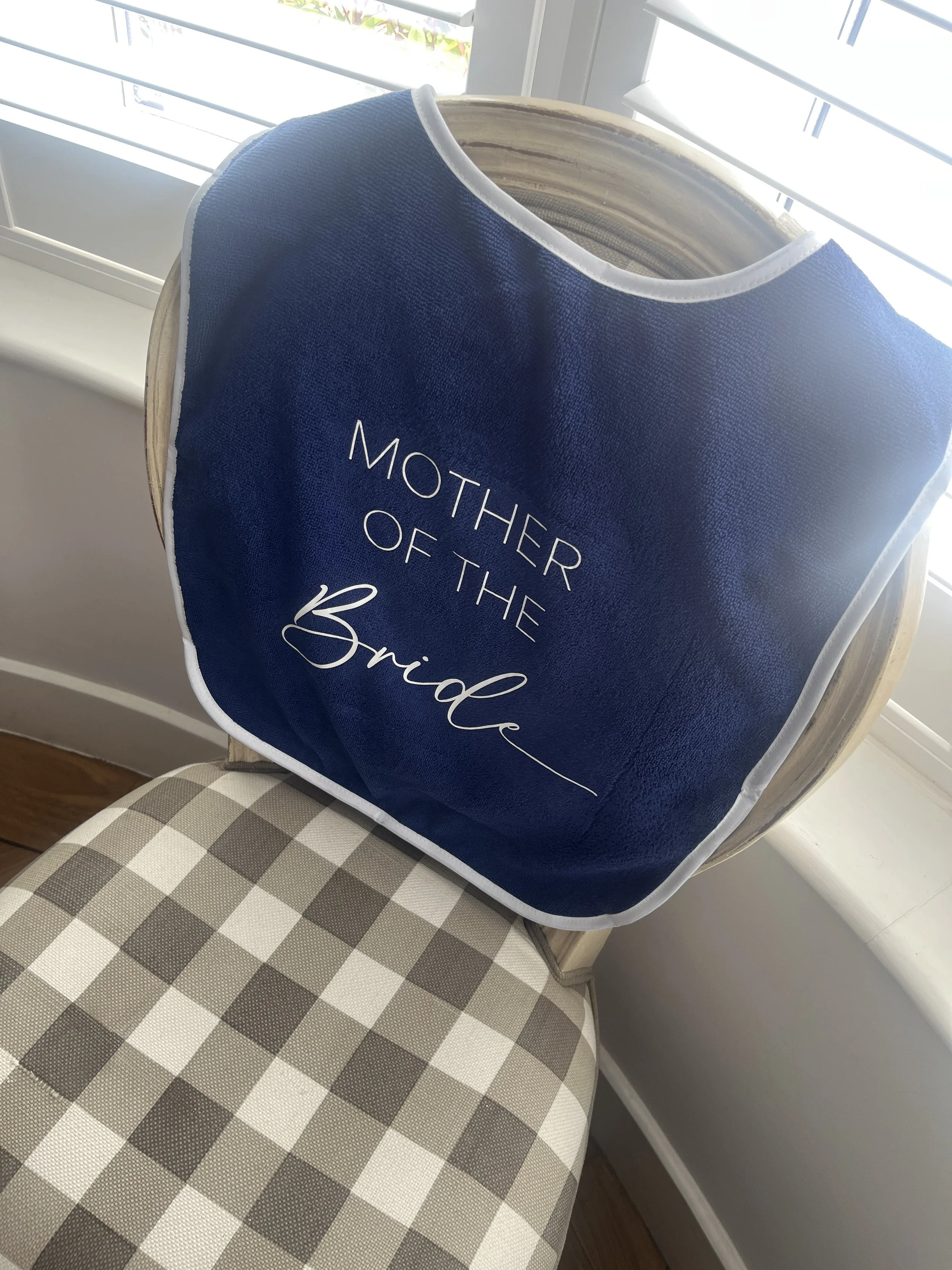 A navy blue fabric cover with white lettering that reads 'Mother of the Bride' placed on a wooden chair near a window with blinds. Personalised merchandise for weddings and events.