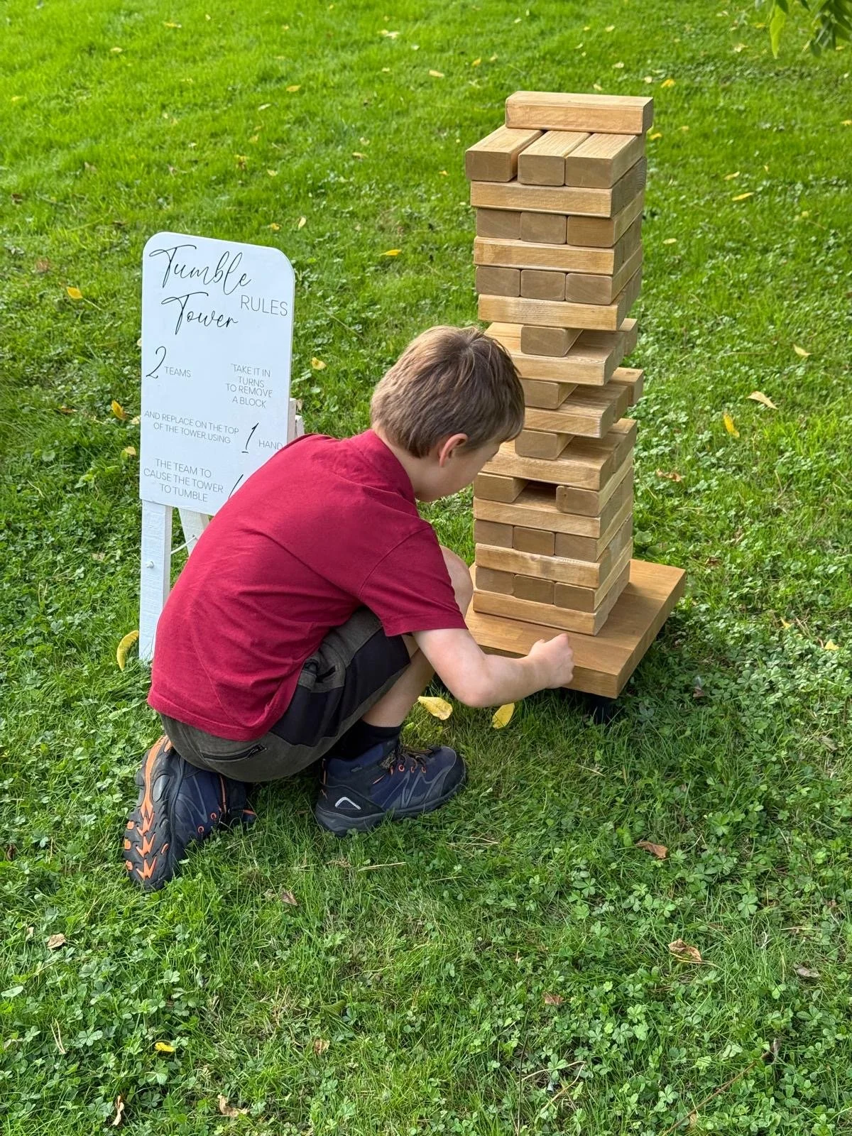 A young boy kneeling on the grass playing a large outdoor Jenga game with wooden blocks. There is a whiteboard with rules for the game titled 'Tumble Tower' next to the game.  Part of a giant lawn games package for hire.