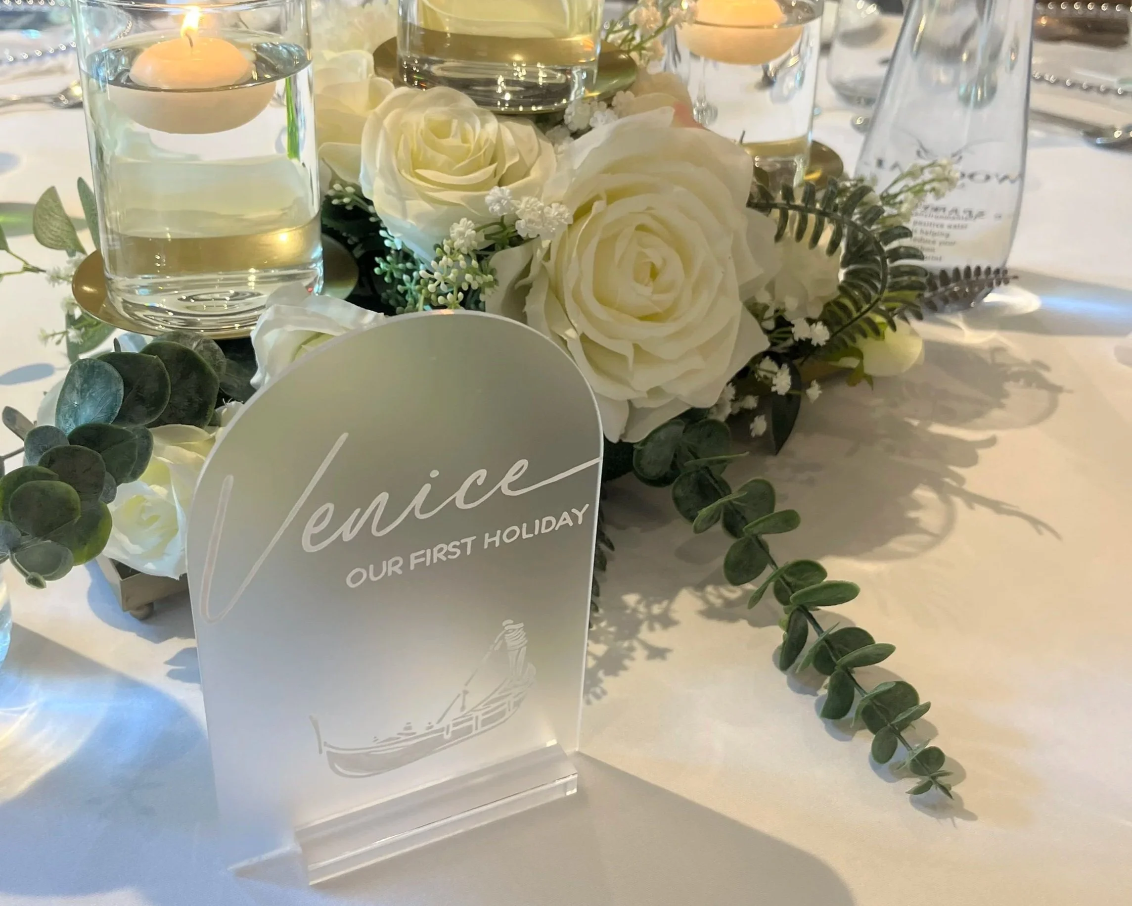 A table setting with a centerpiece of faux white roses, greenery, and small white flowers. Two glass candles with floating candles and water are visible. Personalised event stationery for weddings and events.