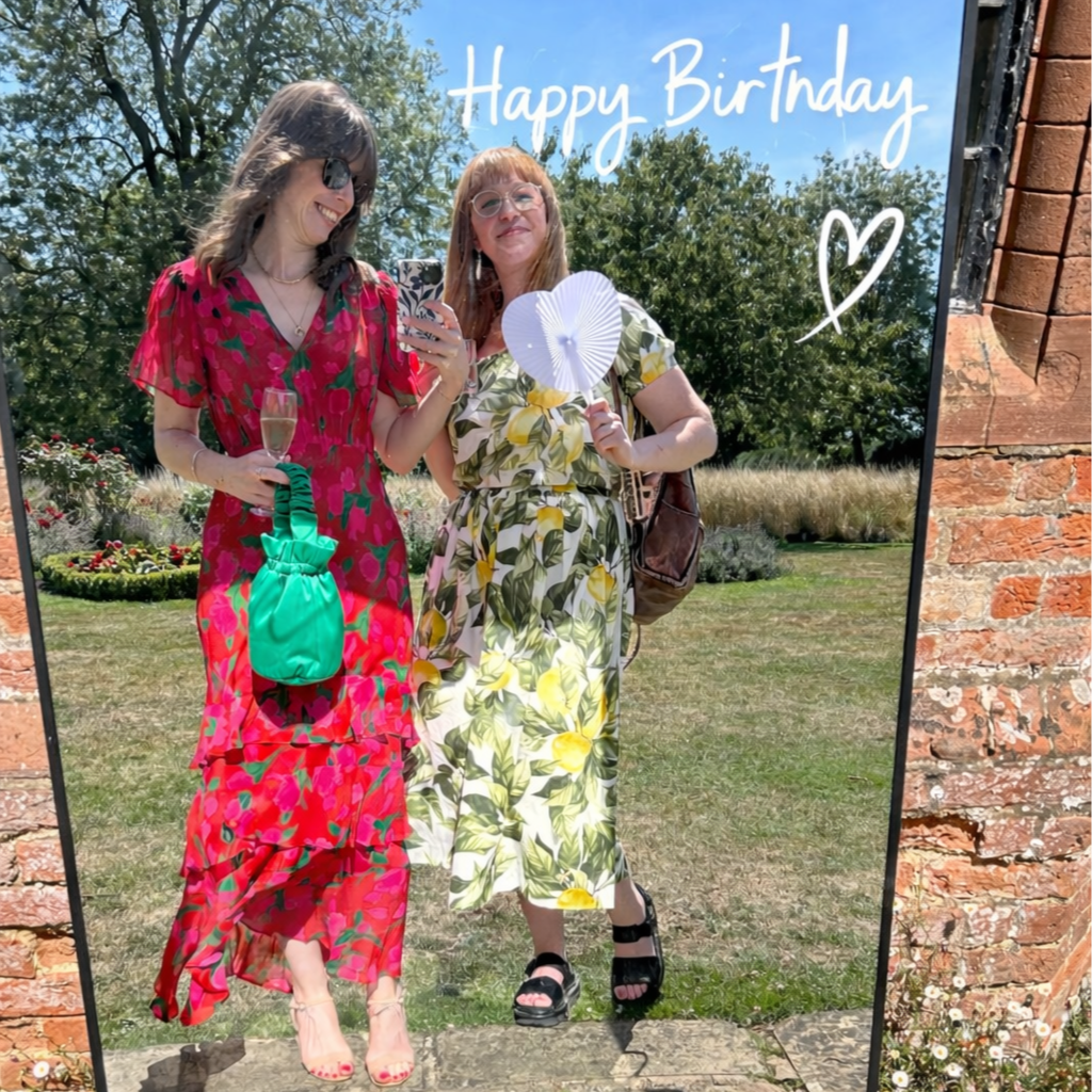Selfie mirror with two women standing in front of a mirror with a 'Happy Birthday' message and a heart, outdoors with trees and grass, celebrating a birthday in summer.