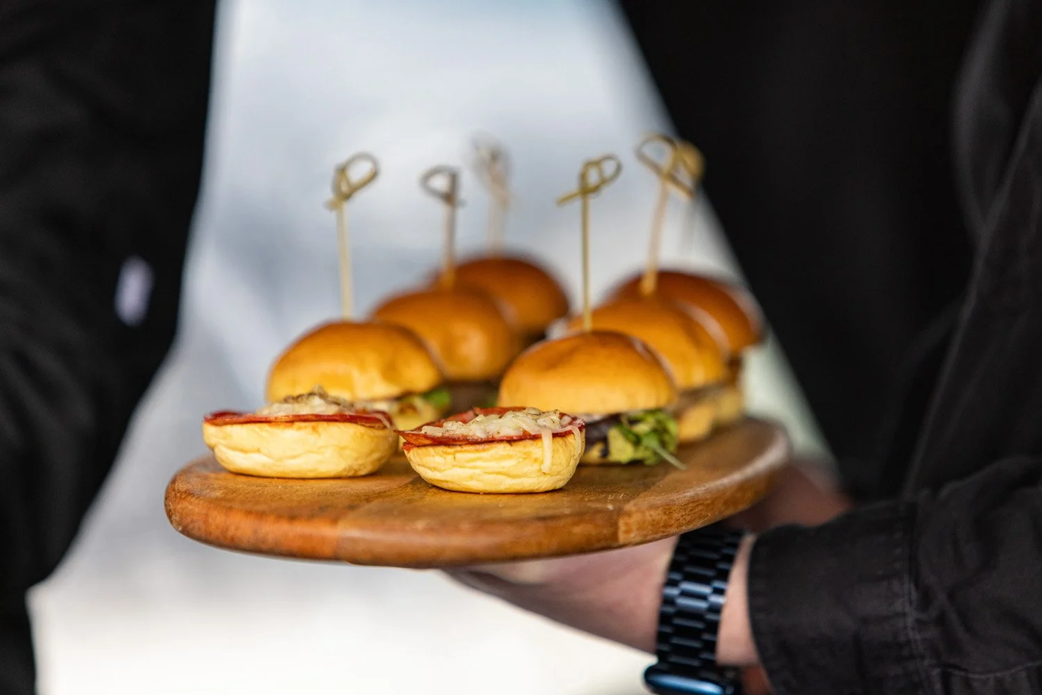 Person holding a wooden tray with several mini sandwiches and sliders, some with crusty buns, some with lettuce and deli meats, with toothpicks in each sandwich. Event planning and catering. 