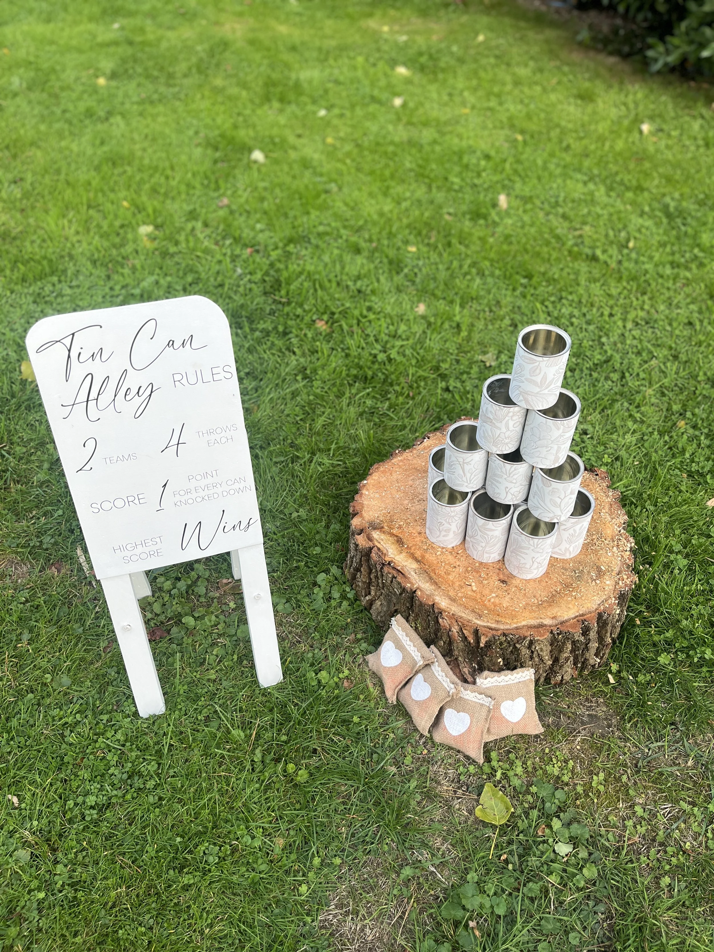 A pyramid of tin cans placed on a tree stump on grass, with a white signboard nearby displaying rules for a game called 'Tin Can Alley'. Forms part of a Giant Lawn Games package.