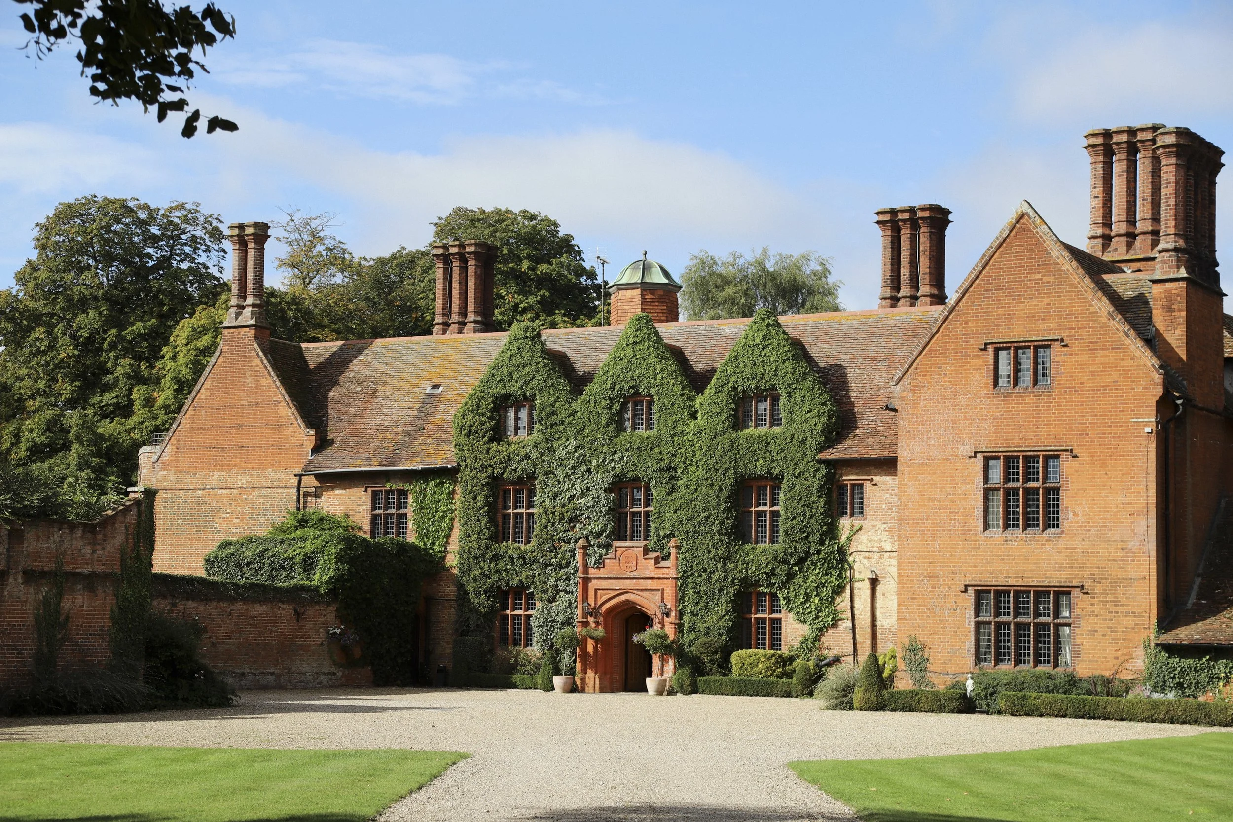 A large brick house with ivy growing across part of its facade, multiple chimneys, and a gravel driveway leading to the entrance.