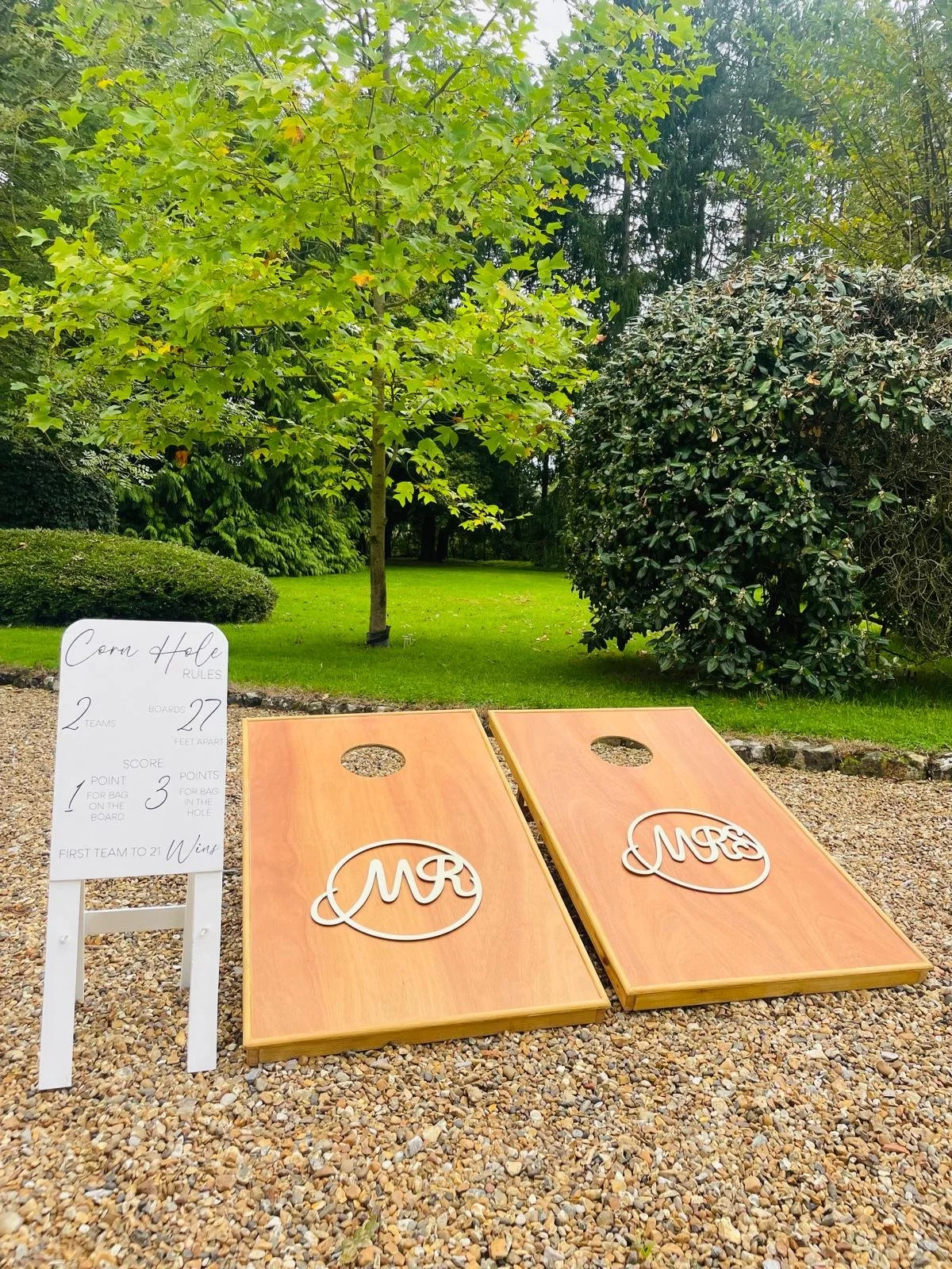 Cornhole game setup outdoors with two wooden boards marked 'Mr' and 'Mrs', set on gravel with trees and bushes in the background, and a whiteboard with game rules. Corn Hole is part of a giant lawn games package for hire.