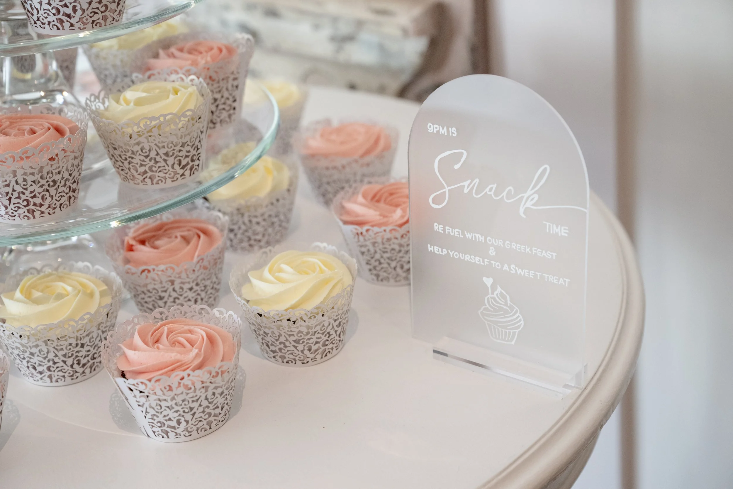 Assorted pastel-colored cupcakes with rose-shaped frosting in white decorative wrappers on a glass tiered stand, with a clear acrylic sign mentioning snack time and Greek feast, on a white table. Personalised event stationery for weddings and events.
