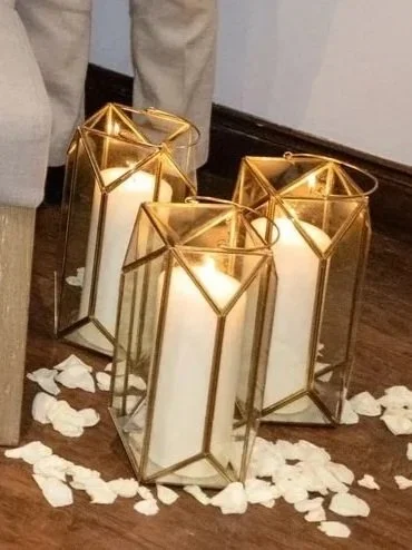 Three large white candles in gold geometric lanterns, surrounded by white flower petals on a wooden floor.