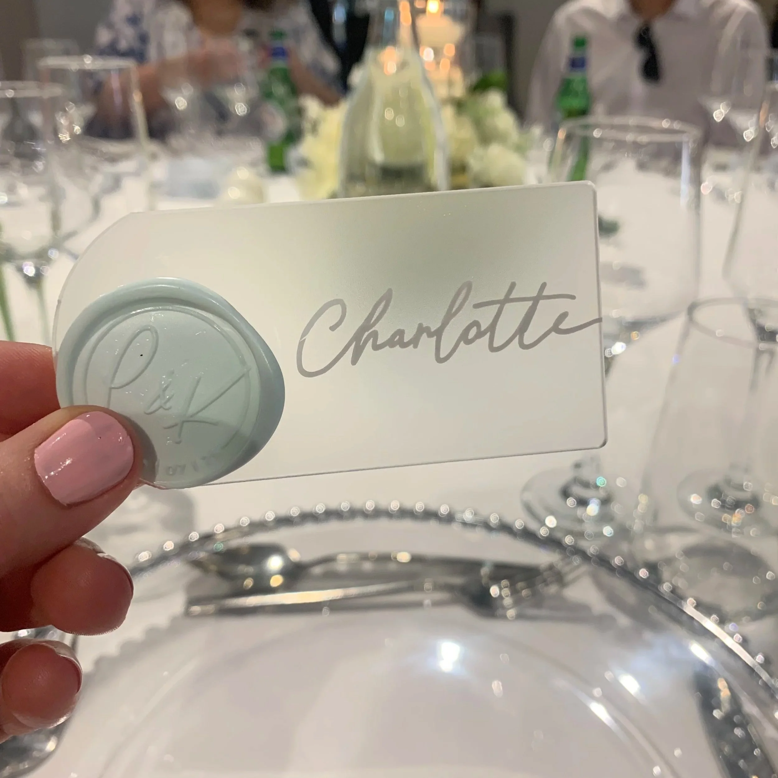 A place card with the name Charlotte, held by a person with pink nails, on a table set for a formal event with glasses, silverware, and a centerpiece in the background. Personalised stationery for weddings and events.
