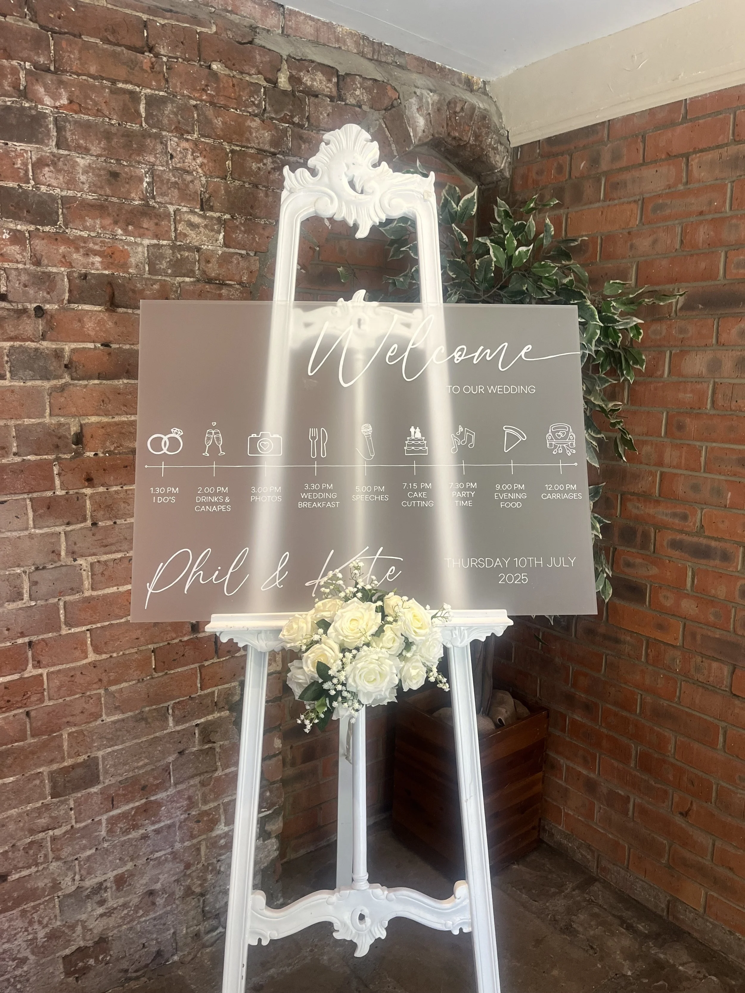 Wedding welcome sign on an easel with schedule and names, decorated with white roses and greenery, against a brick wall. The sign includes a timeline for the wedding day with icons for various events. Personalised wedding and event stationery.